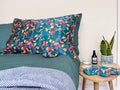 Lifestyle shot of lively hummingbird pillowcase, scrunchie, eye mask and pouch on a bed and table.