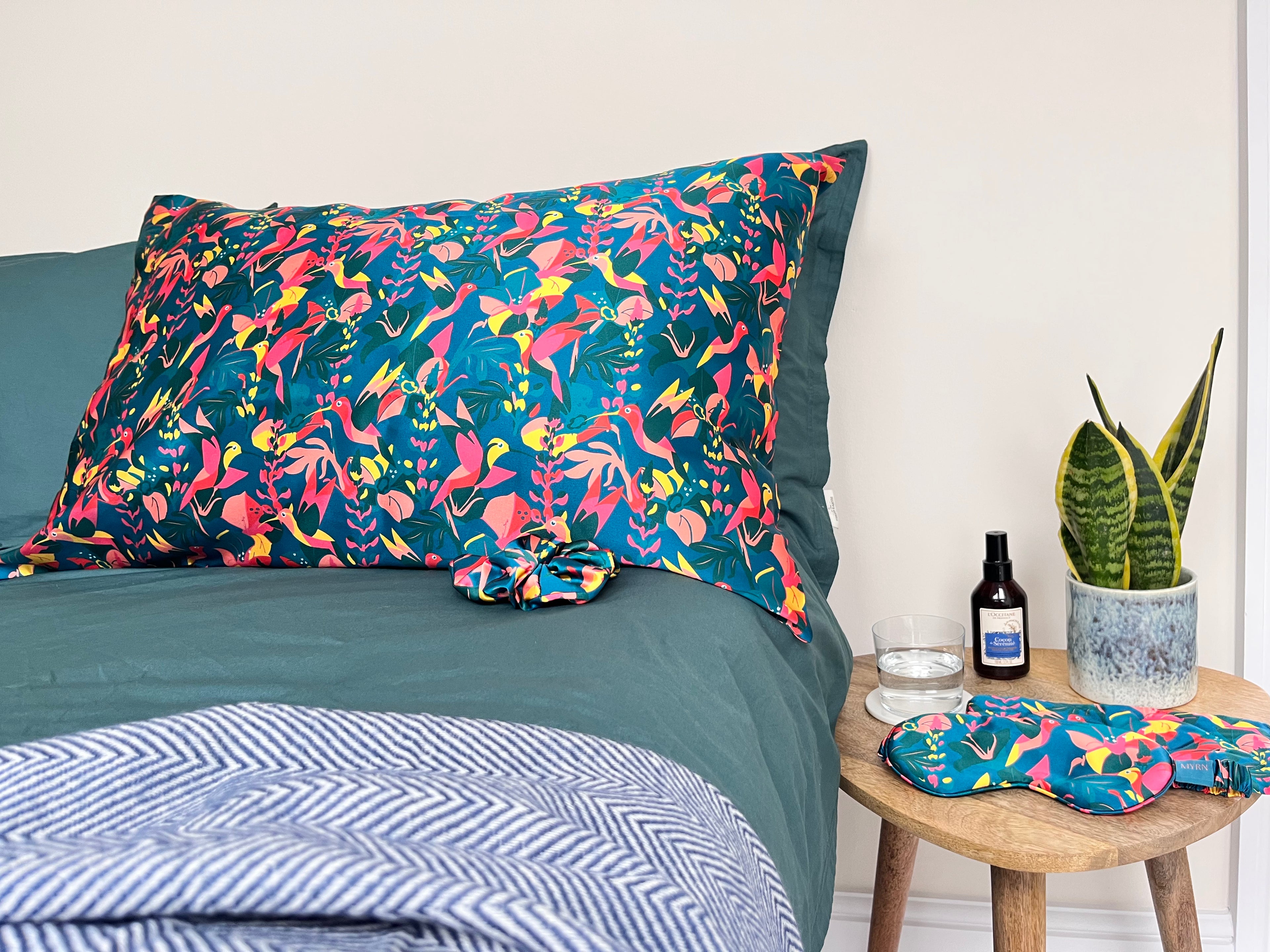 Lifestyle shot of lively hummingbird pillowcase, scrunchie, eye mask and pouch on a bed and table.