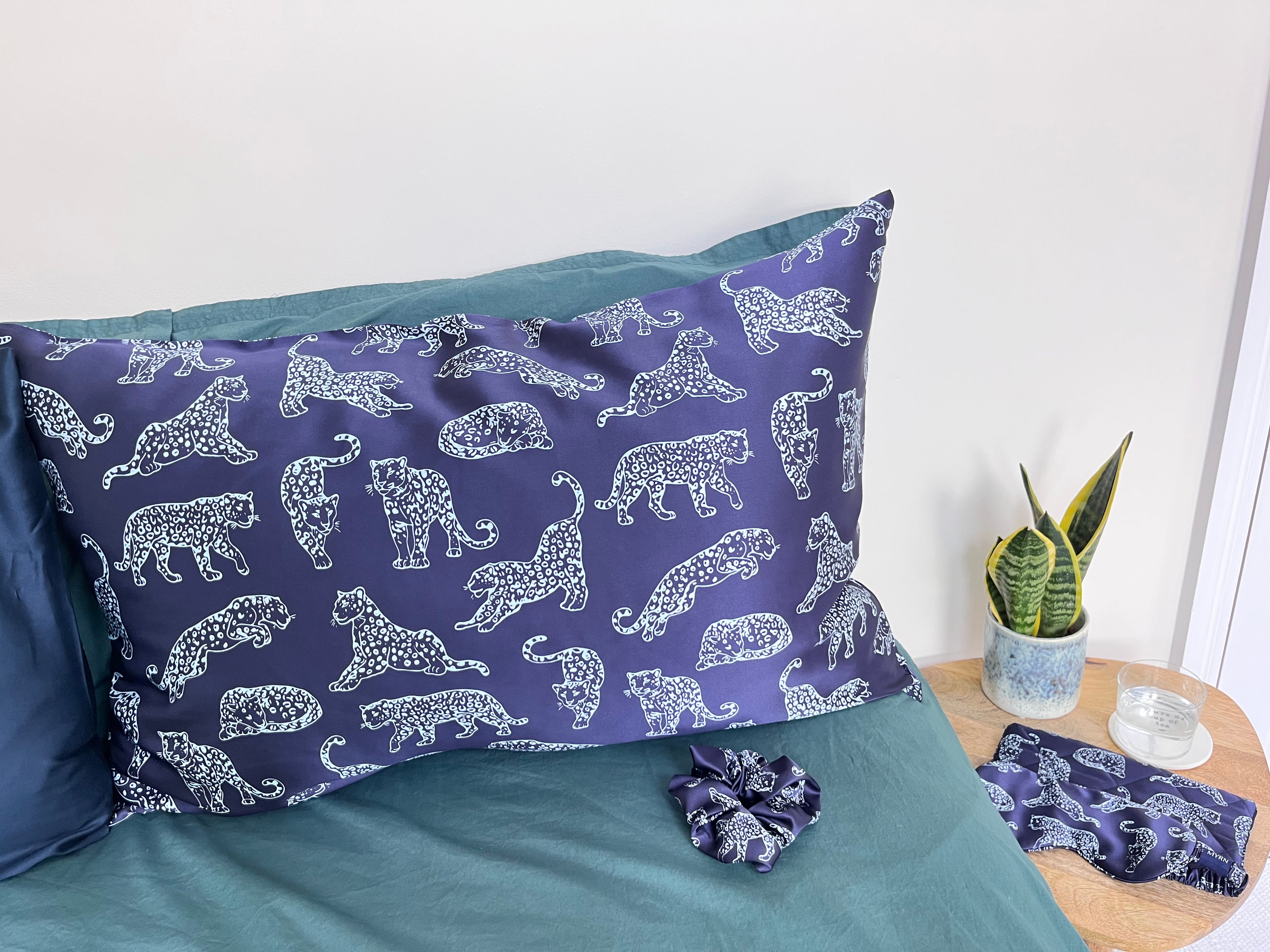 Midnight Leopard silk pillowcase, scrunchie on a bed and eye mask and travel pouch on a bedside table
