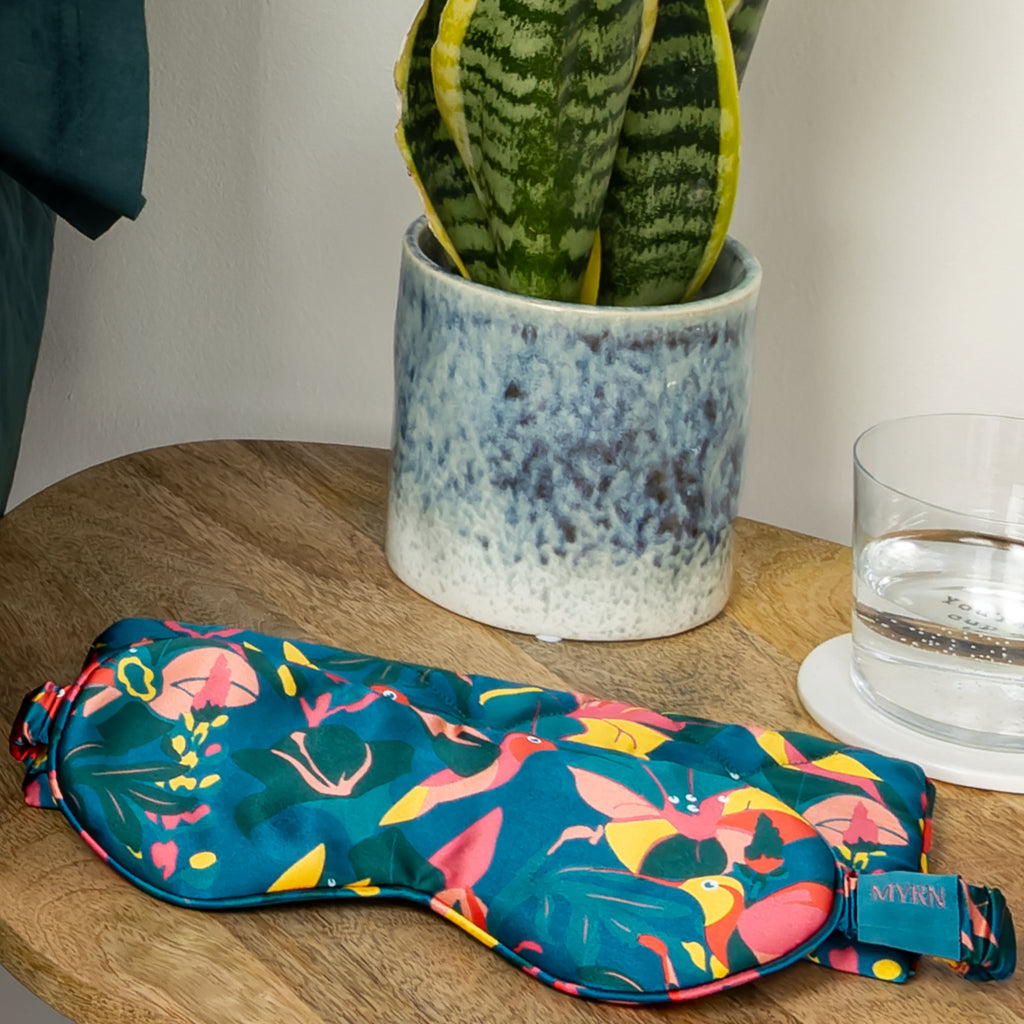 MYRN Silk Hummingbird eye mask and travel case on a bedside table with a sleep plant and glass of water.