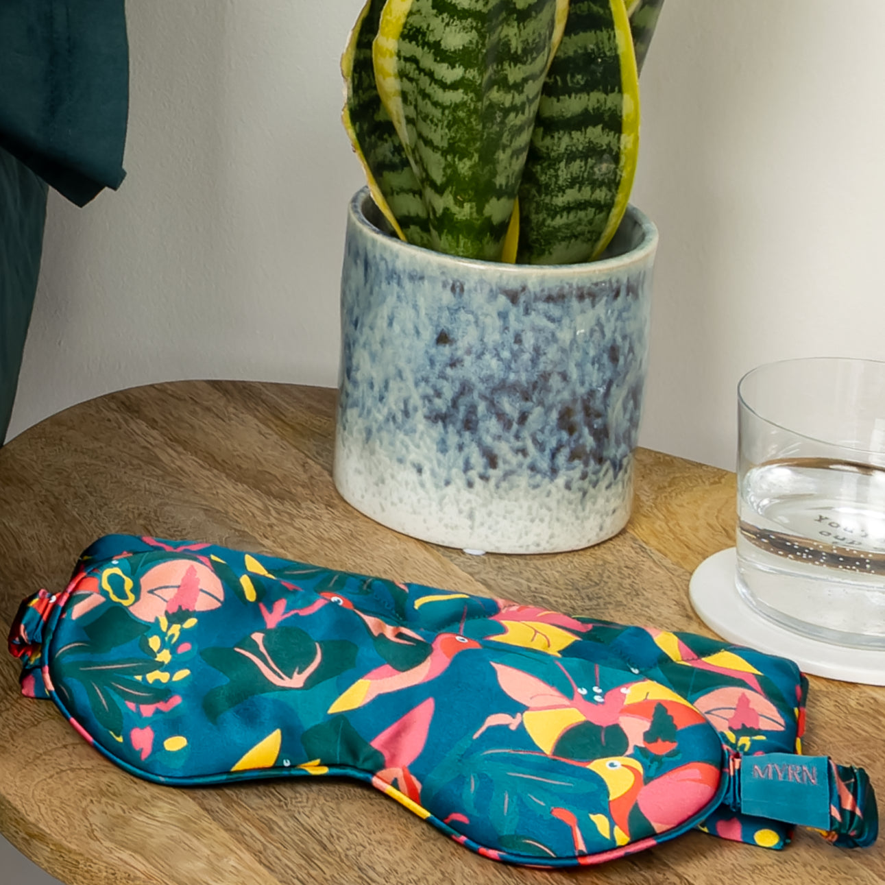 MYRN Silk Hummingbird eye mask and travel case on a bedside table with a sleep plant and glass of water.