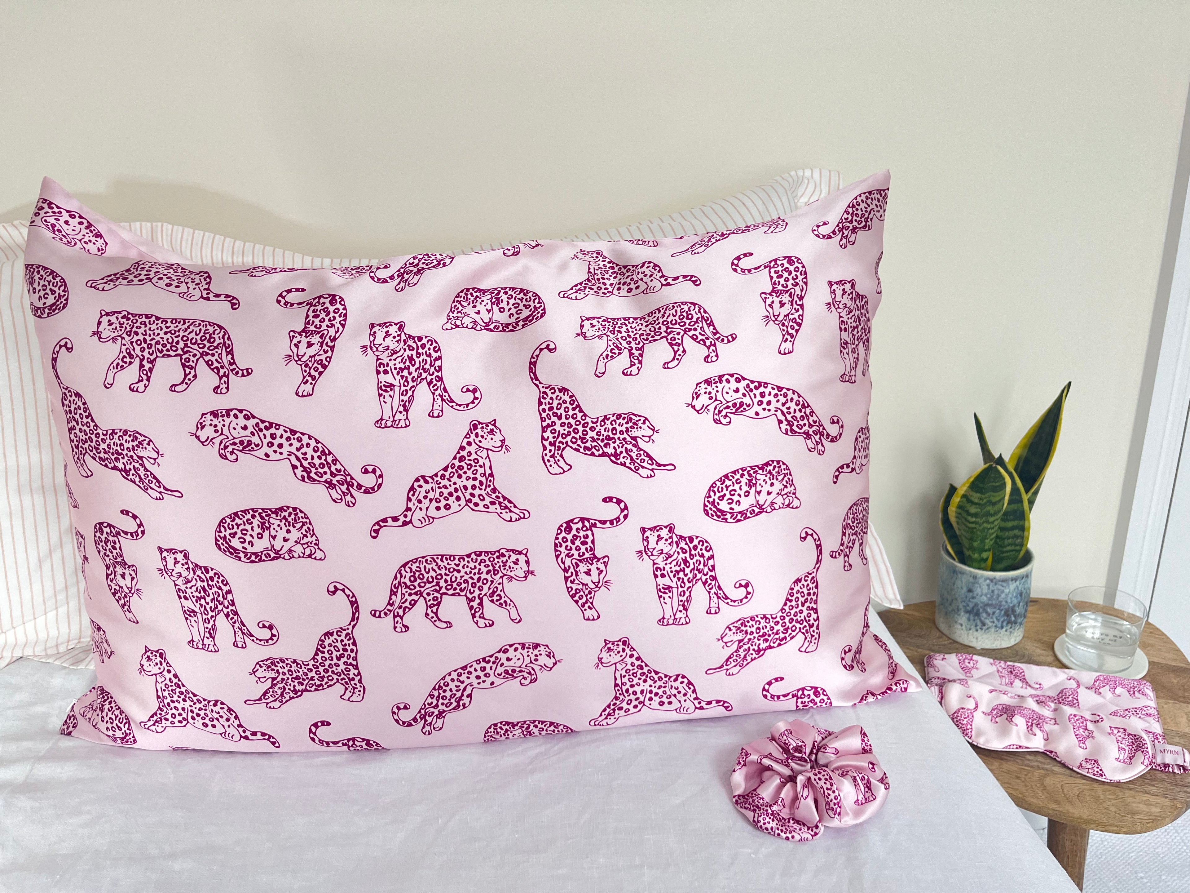 MYRN Silk Pink Leopard printed silk pillowcase and silk scrunchie on a bed and matching eye mask and travel case on a bedside table